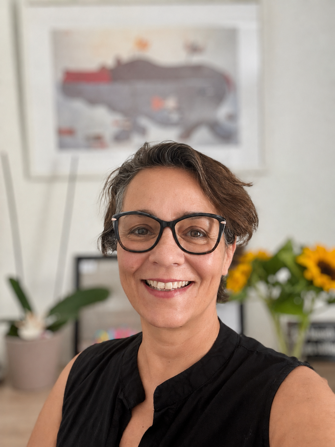Lächelnde Frau mit Brille im Vordergrund, im Hintergrund Blumen und unscharfes Poster an der Wand.